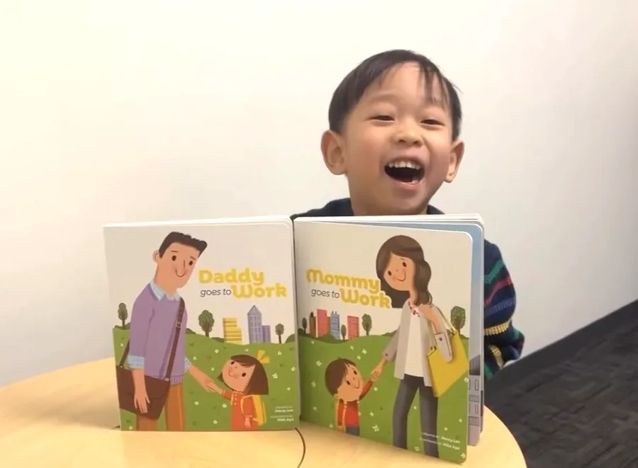 A laughing boy behind copies of Daddy Goes to Work and Mommy Goes to Work