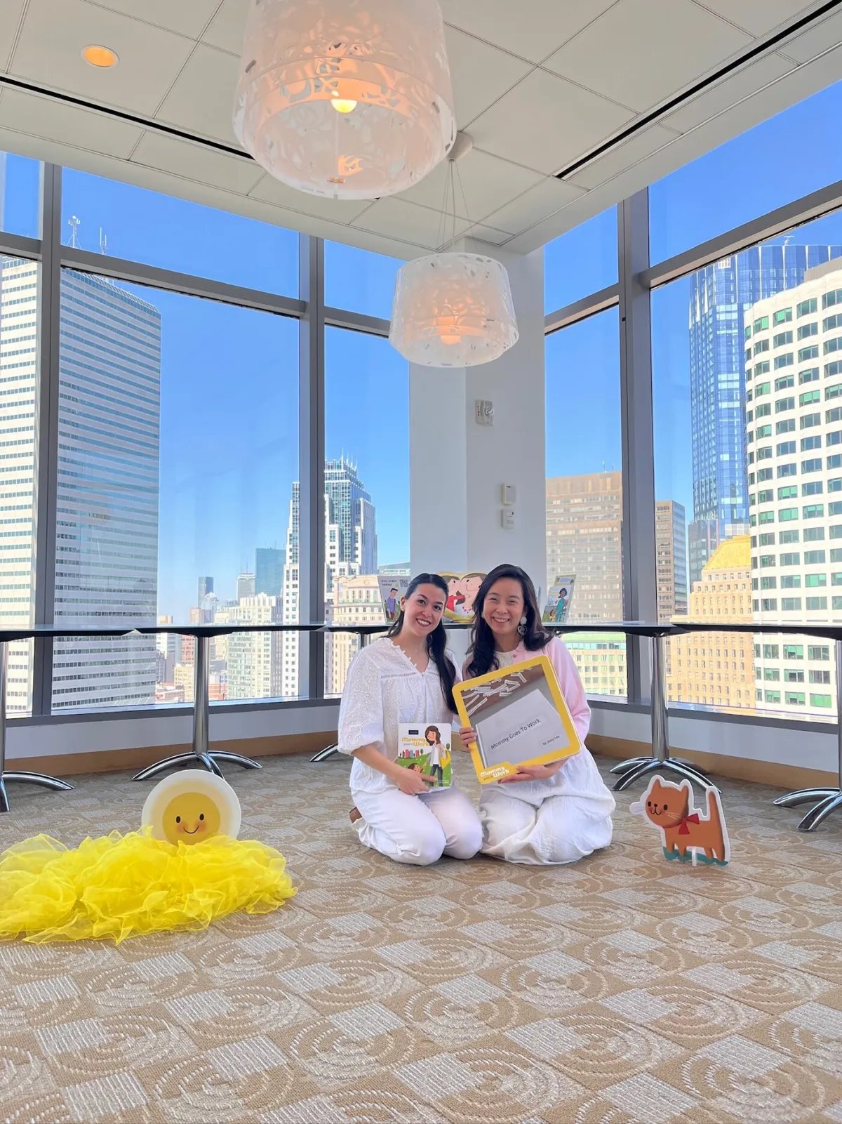 Two facilitators sitting on the floor with Mommy Goes to Work books in an office with city views