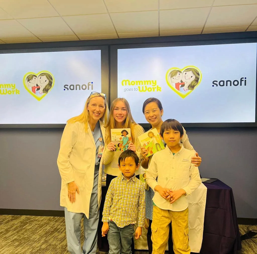 A doctor in a white coat, Jossy Lee and children holding the books at a Mommy Goes to Work event at Sanofi