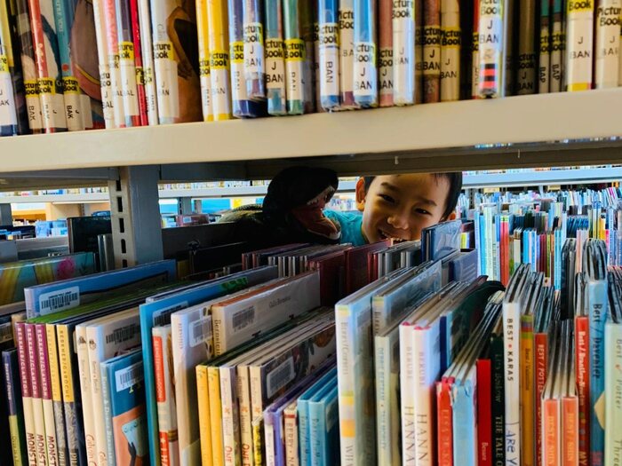 A child peering through a library picture-book shelf, the shelves they searched for the right illustrator