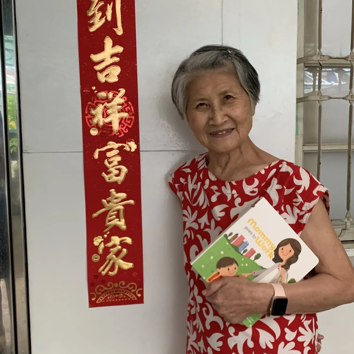 A grandmother holding the book beside a red spring couplet